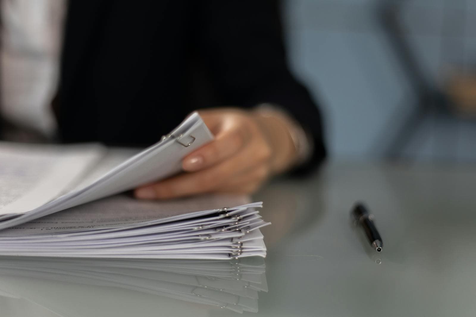 Hands organising a stack of paper documents on a desk beside a pen.